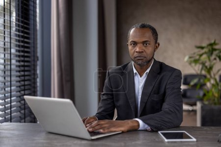 Konzentrierter afroamerikanischer Geschäftsmann im dunklen Anzug arbeitet von seinem Laptop aus in einem hell erleuchteten modernen Büro und strahlt Vertrauen und Sachverstand aus.