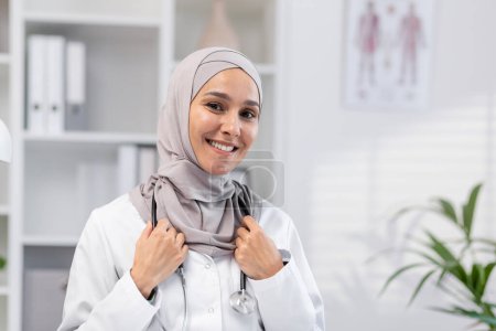 Eine professionelle und freundliche Ärztin im Hidschab, die in ihrem gut beleuchteten Praxiszimmer freundlich lächelt. Stellt Gesundheit, Vielfalt und Professionalität dar.