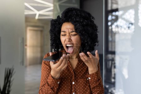African American woman in a polka dot shirt expressing anger and frustration while yelling into a mobile phone in a modern office setting.