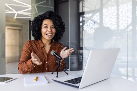 Una alegre mujer afroamericana se involucra con su audiencia en línea mientras graba un podcast en un ambiente de oficina brillante y moderno.
