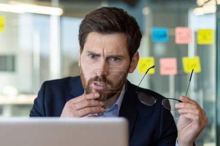 Photo for Focused businessman displays a look of concern while reviewing data on a laptop. He is at a glass-walled office, surrounded by sticky notes. - Royalty Free Image
