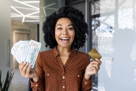 Emocionada mujer afroamericana sosteniendo dinero y una tarjeta de crédito de oro, de pie en un entorno de oficina moderno. Muestra alegría y éxito financiero.