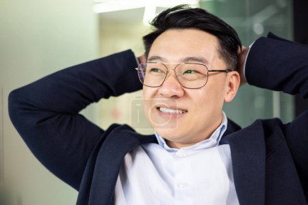 Homme d'affaires asiatique d'âge moyen relaxant avec les mains derrière la tête, souriant dans un environnement de bureau lumineux. Il respire le contentement et la confiance.