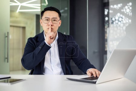 Photo for Asian businessman gesturing for silence with a finger to his lips while seated at a laptop in a stylish, contemporary office environment. - Royalty Free Image