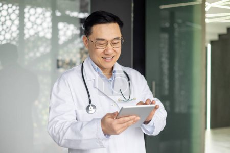 A cheerful Asian male doctor happily interacting with a digital tablet in the bright corridor of a contemporary hospital. Emphasizing professionalism and modern healthcare.