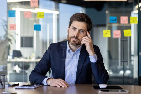 A focused businessman sits in a modern office, contemplating while looking at notes on a glass partition. This professional setting captures a moment of strategic thinking and planning.