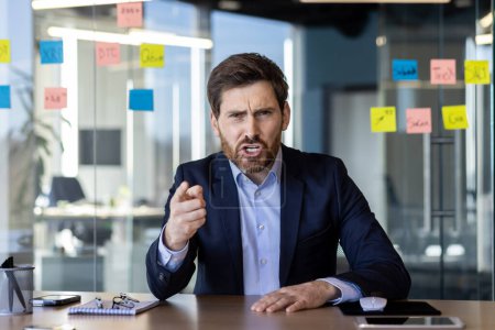 Ein aufgebrachter Geschäftsmann drückt Frustration aus und zeigt in einer hitzigen Diskussion während eines Videotelefonats in einem modernen Büroumfeld.