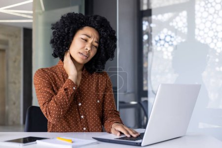 African-American woman experiencing discomfort and neck pain while working on a laptop in a modern office environment, signaling overuse or poor posture.