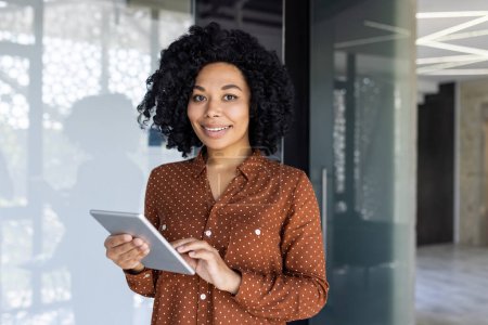 Una mujer afroamericana sonríe con confianza mientras sostiene una tableta en un entorno de oficina moderno, mostrando profesionalismo y facilidad.