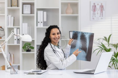 Eine professionelle Radiologin in weißem Laborkittel untersucht und erklärt freudig ein Röntgenbild der Brust in ihrer gut ausgestatteten Praxis, umgeben von medizinischen Diagrammen und einem Laptop..