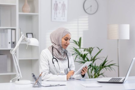 Eine professionelle muslimische Ärztin im Hidschab sitzt in ihrem Büro und benutzt ein digitales Tablet. Das moderne Klinikumfeld stellt eine Mischung aus Technologie und Gesundheitsfürsorge dar.