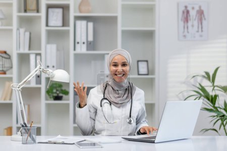 Eine gut gelaunte Ärztin im Hijab grüßt freundlich winkend, während sie an ihrem Schreibtisch in einer hellen, gut organisierten Arztpraxis sitzt. Ihr professionelles Umfeld spiegelt ein modernes, inklusives