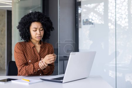A professional African American woman experiences wrist pain while working on her laptop in a modern office setting, reflecting a common work-related issue.