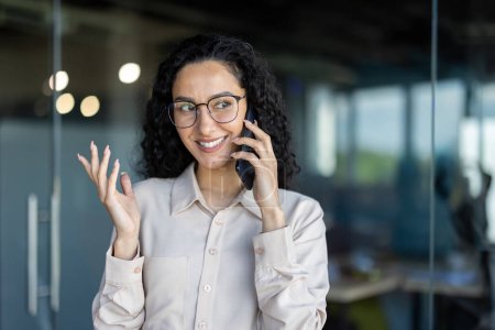 Una mujer profesional sonriente con el pelo rizado habla en un teléfono móvil en un ambiente de oficina contemporáneo, expresando entusiasmo y amabilidad.