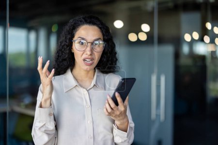 A businesswoman appears upset and frustrated while holding a smartphone in a contemporary office setting. Her expression conveys a sense of urgency and dissatisfaction.