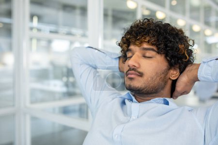 Ein junger Mann in einem hellblauen Hemd mit Knopfdruck entspannt sich mit geschlossenen Augen und streckt seinen Hals, während er in einem hellen, modernen Büroraum sitzt und ein Gefühl von Leichtigkeit und Frieden ausstrahlt..