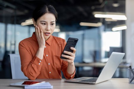 Femme d'affaires asiatique concentrée en chemisier orange regardant perplexe tout en tenant un smartphone à son espace de travail de bureau moderne. Elle semble réagir à des nouvelles inattendues.