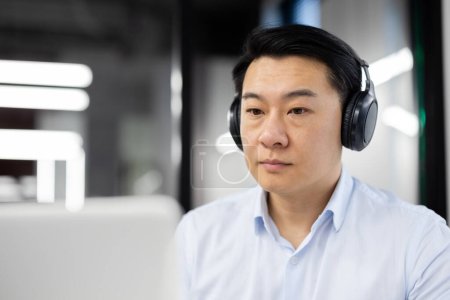 Ein professioneller asiatischer Mann in Businesskleidung konzentriert sich, während er Kopfhörer trägt. Er wirkt engagiert und seriös, sitzt in einem modernen Büro.