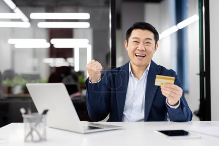 Téléchargez les photos : Joyeux homme d'affaires asiatique en costume détient une carte de crédit, célébrant le succès financier à son bureau entouré de gadgets modernes. - en image libre de droit