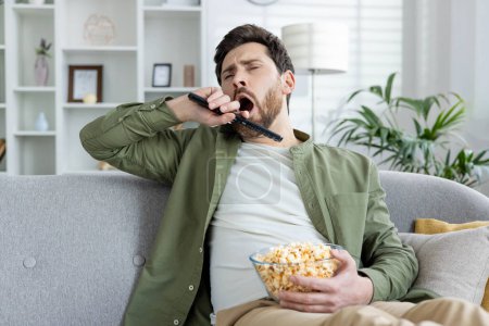 Photo for A bored young man yawning while holding a TV remote and a bowl of popcorn, sitting on a sofa in a living room setting. - Royalty Free Image