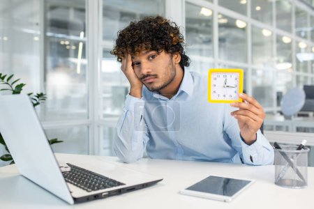 Ein junger Mann sitzt an seinem Schreibtisch im Büro und zeigt Anzeichen von Langeweile und mangelnder Motivation, während er eine Uhr hält, die den langsamen Lauf der Zeit signalisiert..