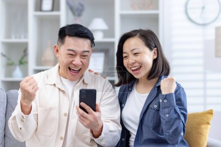 An Asian couple is visibly thrilled as they celebrate a victory, looking at a smartphone together in a cozy living room. Their joyful expressions and enthusiastic gestures highlight a happy