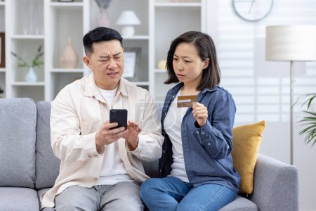 Asian couple looks distressed as they uncover unauthorized charges on their credit card phone, man confusion while the woman, holding the card, seems anxious. stress, frustration of financial fraud.