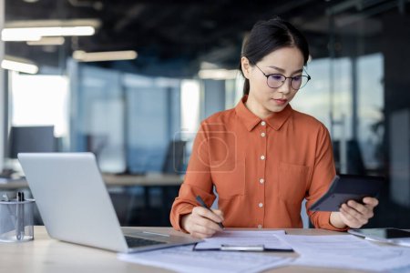 Sérieuse jeune femme d'affaires asiatique axée sur l'audit de documents financiers, tenant une calculatrice dans un cadre de bureau moderne.