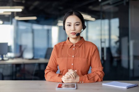 A focused professional Asian woman, wearing an orange shirt and headset, engages in a serious video call, looking directly at the camera in a modern office setting.