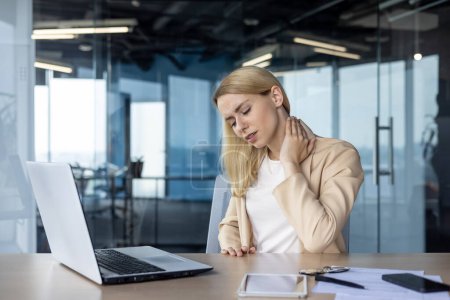 Junge Geschäftsfrau spürt Nackenschmerzen, während sie am Schreibtisch am Laptop arbeitet. Konzept von arbeitsbedingtem Stress und Unbehagen.