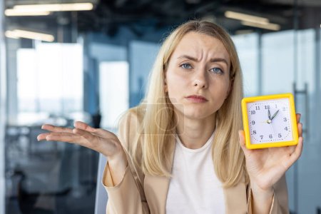 A confused businesswoman holds a yellow clock in a modern office, expressing uncertainty about time management and punctuality.