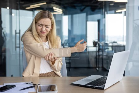 Eine junge professionelle Frau erlebt während der Laptopnutzung in einem modernen Büroumfeld Beschwerden in ihrem Arm, die auf eine mögliche wiederholte Belastung hindeuten..