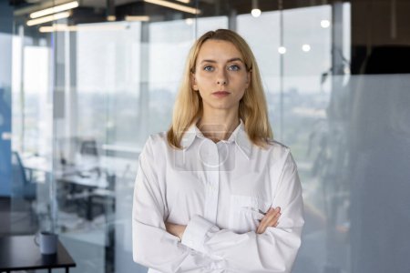 A professional blond businesswoman stands confidently with arms crossed in a modern office setting, exuding competence and authority.