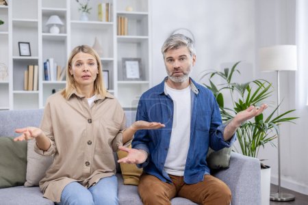 Middle-aged couple sitting on the sofa with confused expressions in a modern living room, conveying feelings of uncertainty and misunderstanding