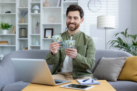 Hombre sonriente contando dinero en casa mientras utiliza el ordenador portátil para el éxito financiero y la inversión. Concepto de ganar dinero en línea, trabajo remoto y planificación financiera.