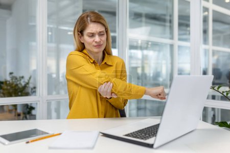 A woman in a yellow shirt experiencing elbow pain while working on her laptop in a modern office environment.