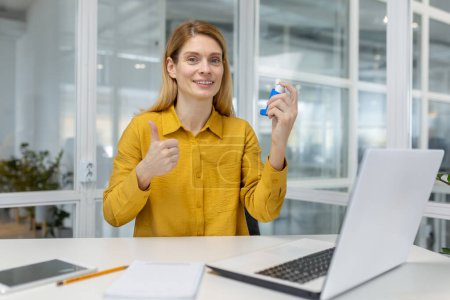 Smiling woman using an asthma inhaler and giving a thumbs-up sign while seated at desk in a modern office environment.
