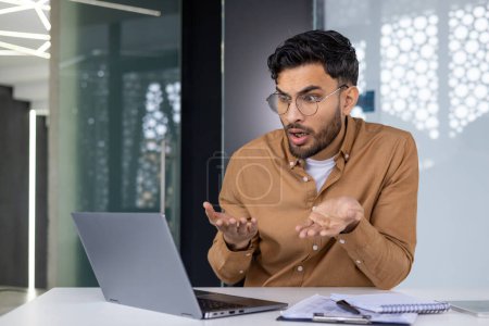 Un homme confus portant des gestes de lunettes à l'écran de son ordinateur portable tout en travaillant dans un bureau moderne, exprimant frustration ou malentendu.