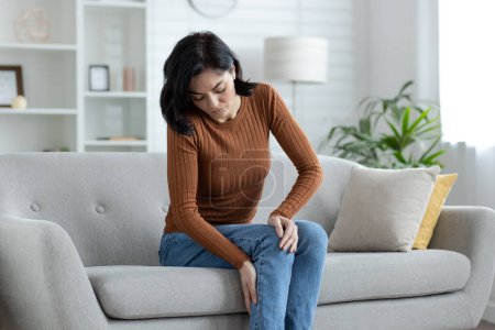 Une femme en tenue décontractée assise sur un canapé à la maison, souffrant de douleurs au genou. L'image capture l'inconfort et les problèmes de santé.
