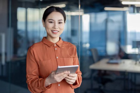 Confident Asian businesswoman smiling while holding a tablet in a modern office setting with glass walls.