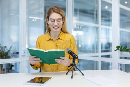 Foto de Woman in yellow shirt reading a book aloud in a modern office, using a microphone, with a tablet and notebook on the table. - Imagen libre de derechos