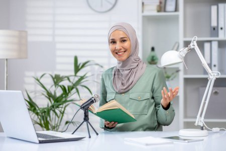 Confident woman in hijab hosting a podcast at her home desk. Holding a book in front of a microphone while smiling towards the camera.