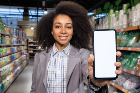 In a supermarket aisle, a young African American woman is displaying a smartphone with a blank screen amidst various products. This showcases the blend of technology and shopping experience