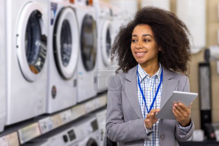 In an appliance store, a happy woman with curly hair smiles holding a tablet. She is comparing options in front of washing machines and dryers, possibly making a purchase or gathering information for