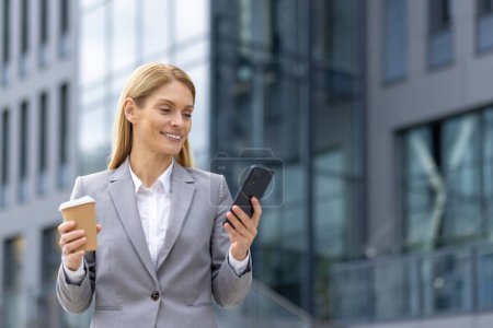 In a modern city, a professional businesswoman in a grey suit smiles while checking her phone and holding a coffee cup outside an office building. She exudes confidence and success