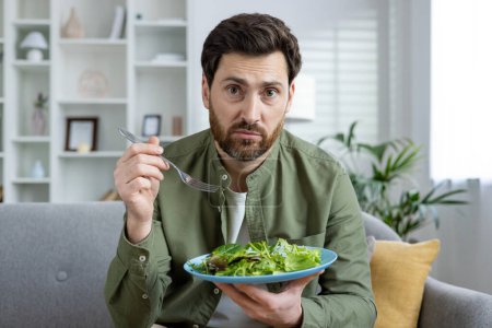 Confused man sitting at home holding a plate of salad with a fork, expressing uncertainty about his meal.