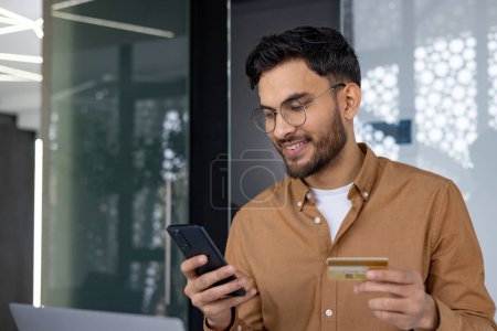 Téléchargez les photos : Homme souriant utilisant son smartphone et sa carte de crédit pour effectuer un paiement en ligne dans un cadre de bureau moderne. - en image libre de droit