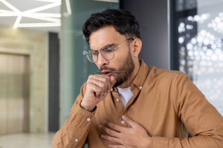 A young man coughing with hand on chest in a modern office environment, indicating illness or discomfort.