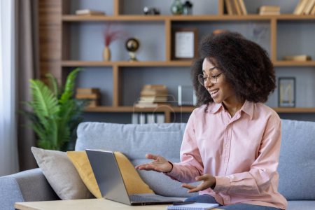 A woman with curly hair is smiling on a couch as she works on her laptop, surrounded by shelves with books and decor. This cozy and productive home office offers a comfortable workspace