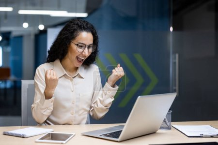 Femme aux cheveux bouclés avec des lunettes célèbre le succès du travail dans un bureau moderne, levant les poings joyeusement tout en regardant son ordinateur portable, reflétant la réalisation et la joie dans un cadre professionnel